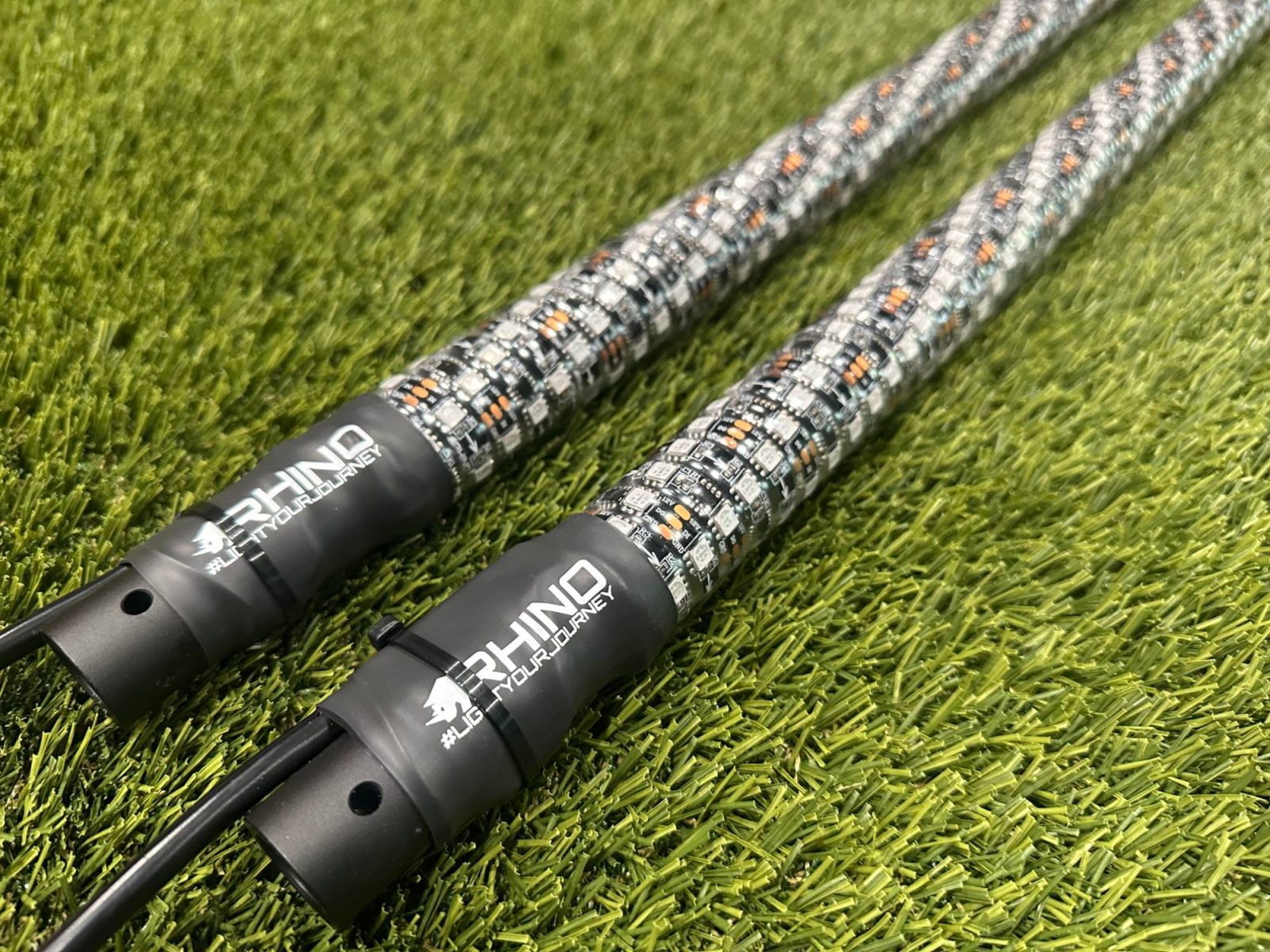 Two Rhino 2.0 whips lying on turf, branded collars toward the camera, the wrapped LED strip running away out of frame